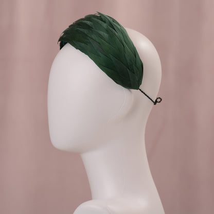 A dark green feather headpiece in a wide headband style. The feathers are cut to points and create texture and depth as they wrap around the head. The hat is fitted with a hat elastic. It is displayed on a white mannequin head against a soft pink background.