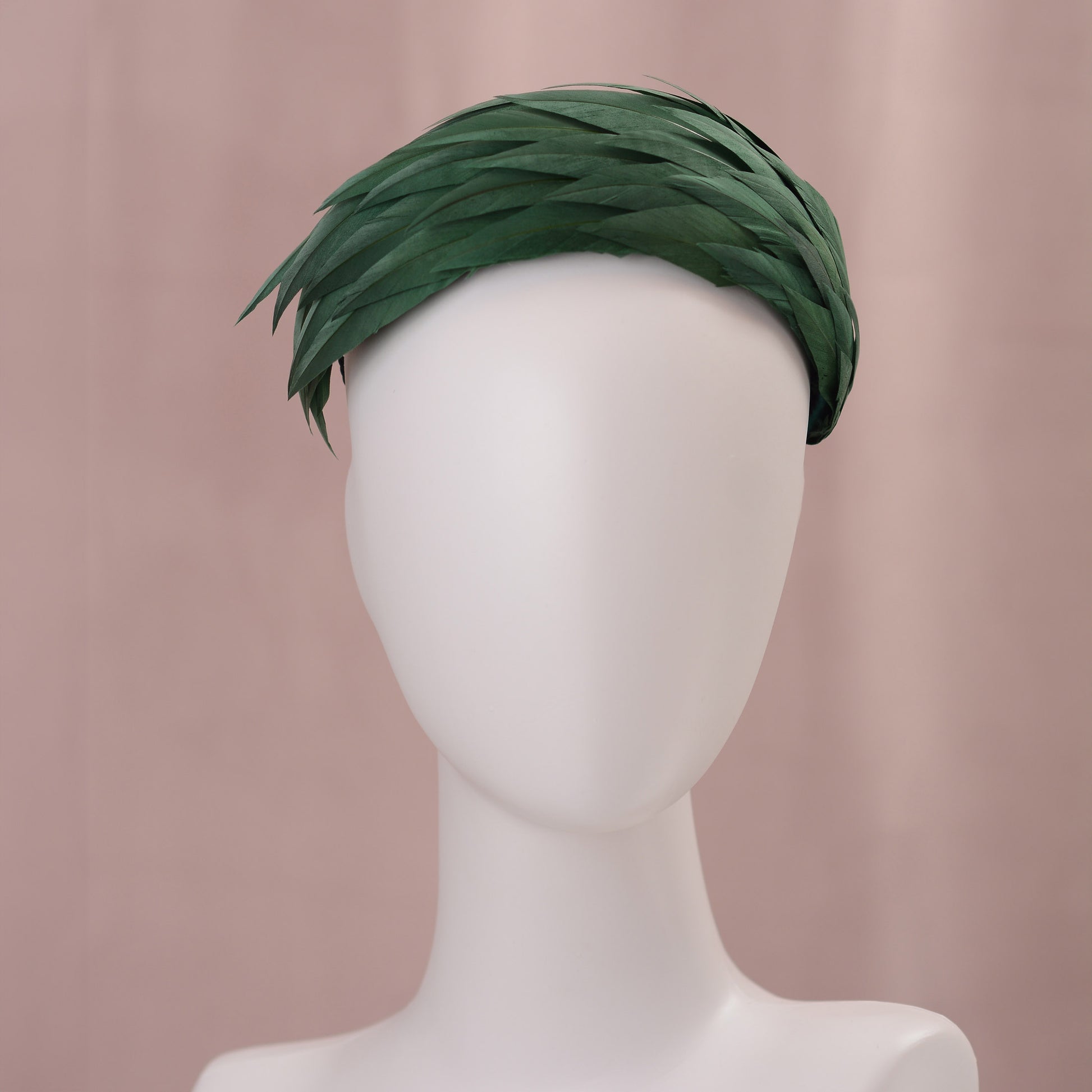 A dark green feather headpiece in a wide headband style. The feathers are cut to points and create texture and depth as they wrap around the head. It is displayed on a white mannequin head against a soft pink background.