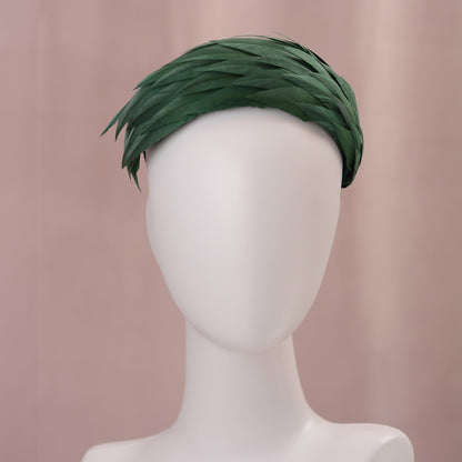 A dark green feather headpiece in a wide headband style. The feathers are cut to points and create texture and depth as they wrap around the head. It is displayed on a white mannequin head against a soft pink background.
