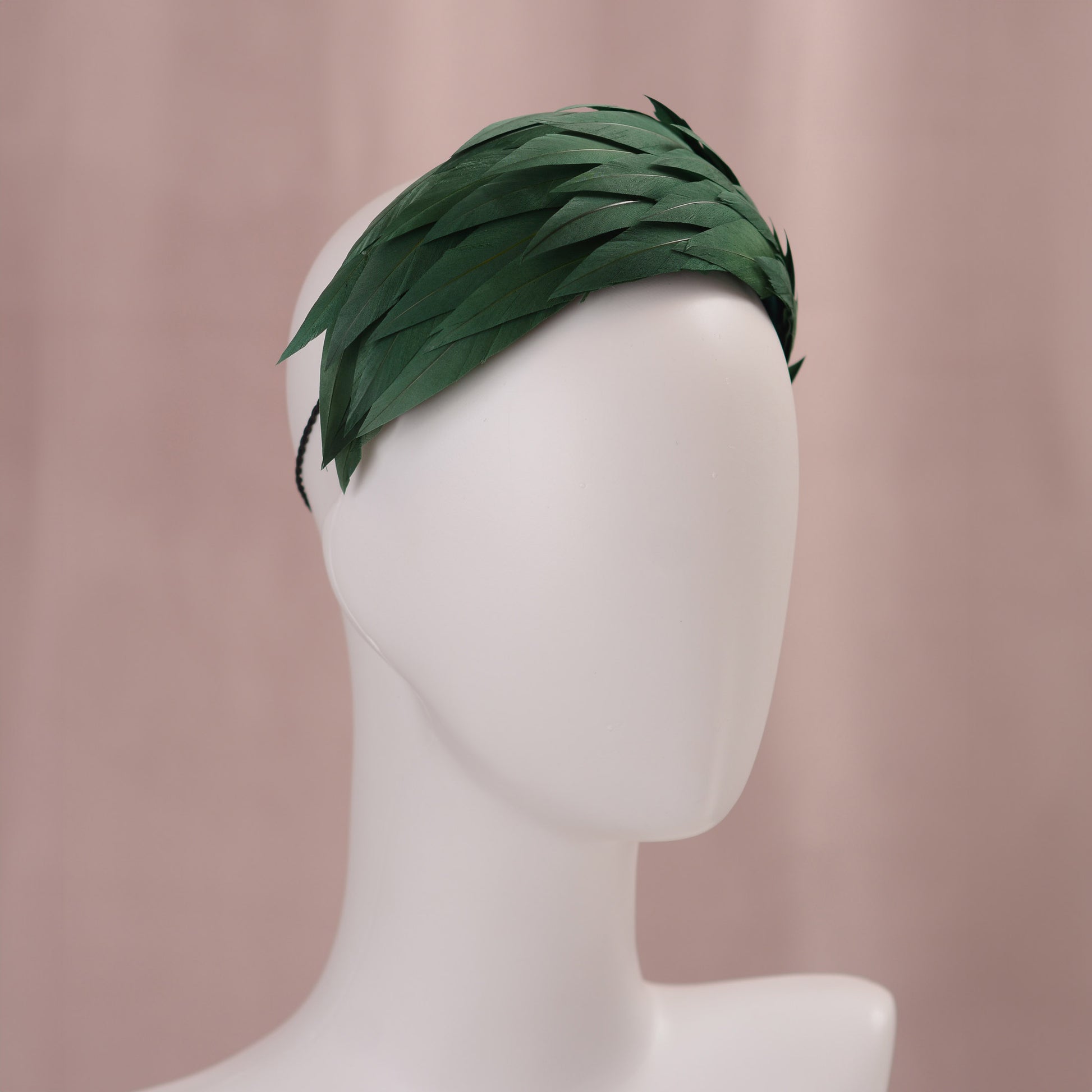 A dark green feather fascinator in a wide headband style. The feathers are cut to points and create texture and depth as they wrap around the head. The hat is fitted with a hat elastic. It is displayed on a white mannequin head against a soft pink background.