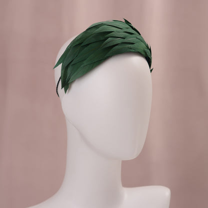 A dark green feather fascinator in a wide headband style. The feathers are cut to points and create texture and depth as they wrap around the head. The hat is fitted with a hat elastic. It is displayed on a white mannequin head against a soft pink background.