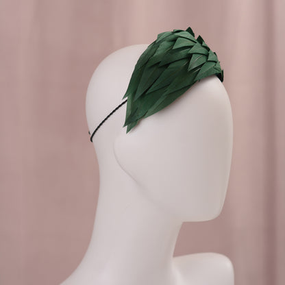 A dark green feather headpiece in a wide headband style. The feathers are cut to points and create texture and depth as they wrap around the head. The hat is fitted with a hat elastic. It is displayed on a white mannequin head against a soft pink background.