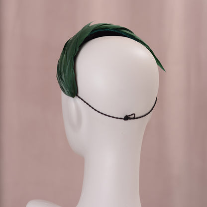 A dark green feather headpiece in a wide headband style. The feathers are cut to points and create texture and depth as they wrap around the head. The hat is fitted with a hat elastic. It is displayed on a white mannequin head against a soft pink background.