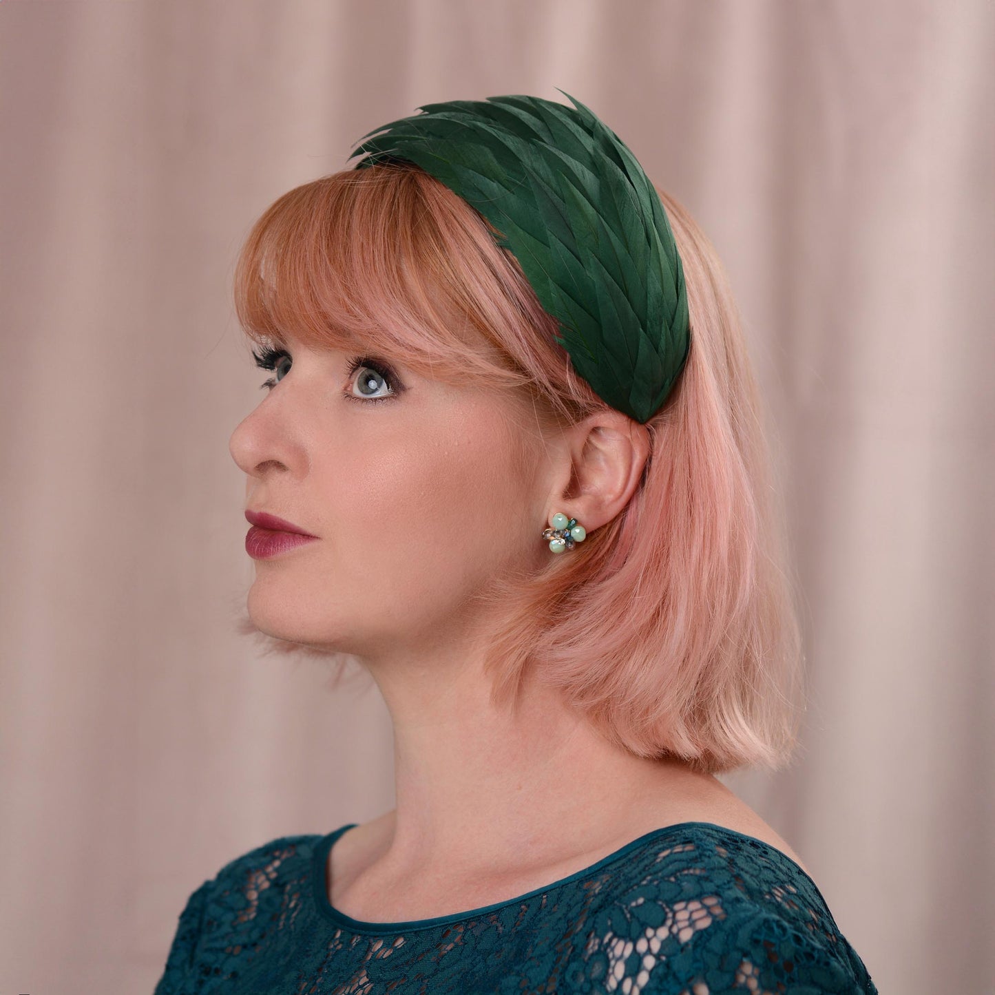 A woman with short pink hair wearing a dark green feather  wedding fascinator in a wide headband shape, photographed against a soft beige curtain background.