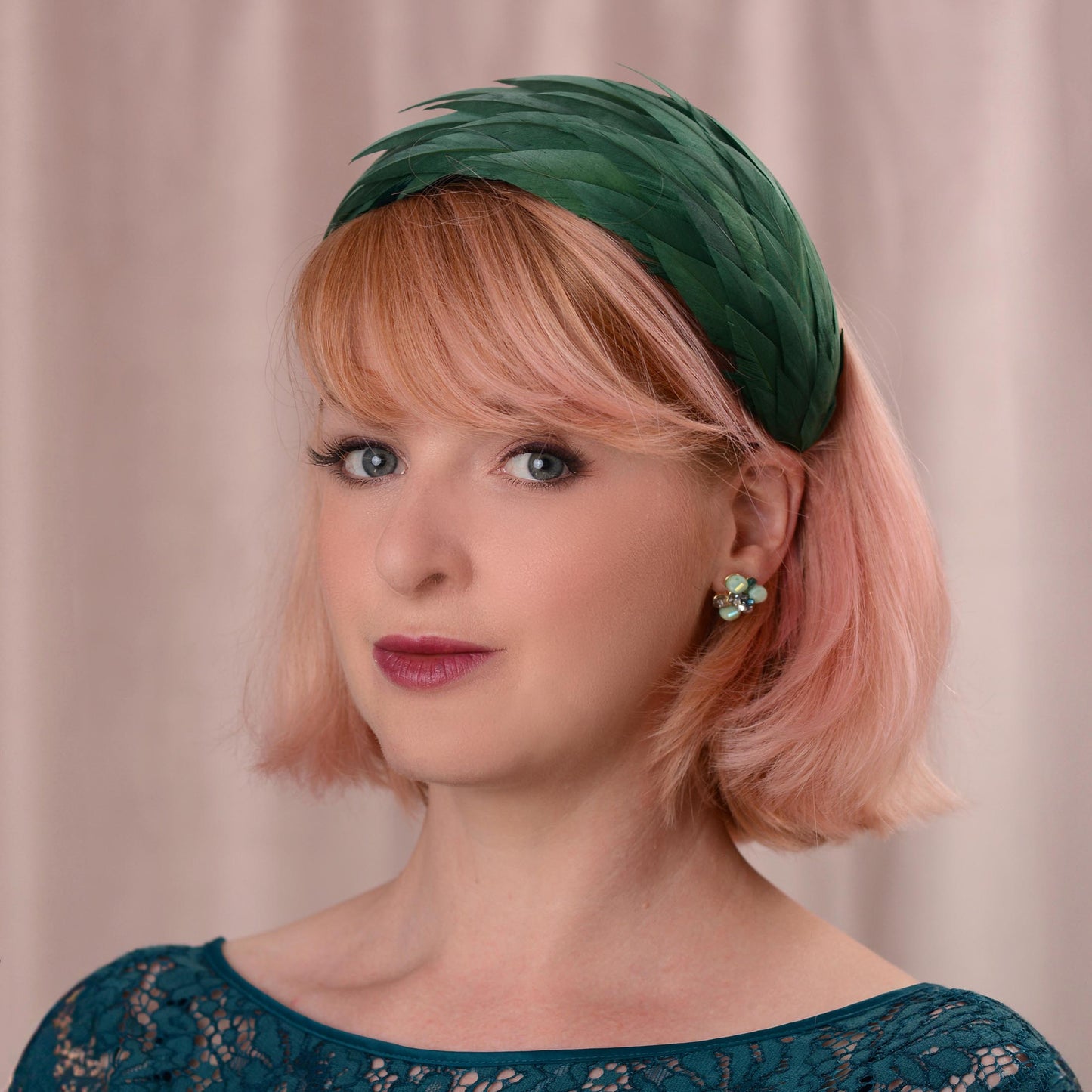 A woman with short pink hair wearing a dark green feather fascinator in a wide headband shape, photographed against a soft beige curtain background.