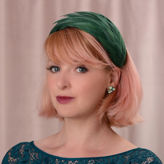 A woman with short pink hair wearing a dark green feather fascinator in a wide headband shape, photographed against a soft beige curtain background.