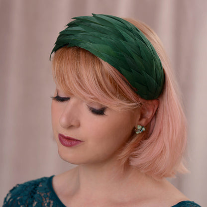 A woman with short pink hair wearing a dark green feather  horse racing fascinator in a wide headband shape, photographed against a soft beige curtain background.