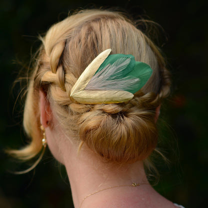 A hair clip with green, pink, and gold feathers adorning a woman with blonde hair in a braided up-do.