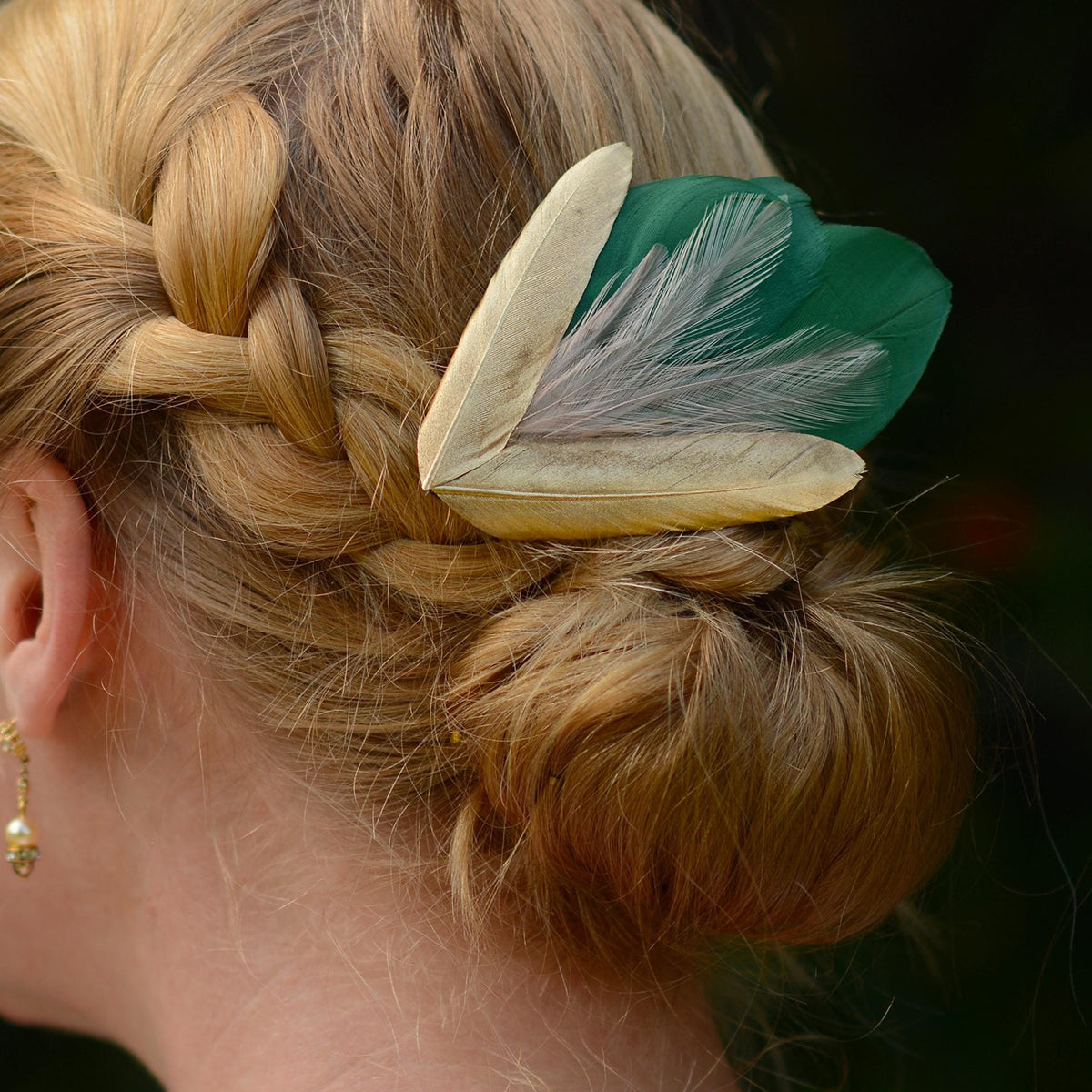 A close-up of a woman's braided and bun hairstyle featuring a decorative hair accessory with two green feathers layered with wispy blush pink feathers andtwo gold feathers in a V-shape at the bottom.
