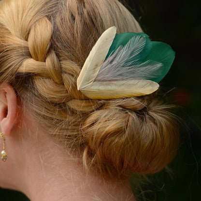 A close-up of a woman's braided and bun hairstyle featuring a decorative hair accessory with two green feathers layered with wispy blush pink feathers andtwo gold feathers in a V-shape at the bottom.