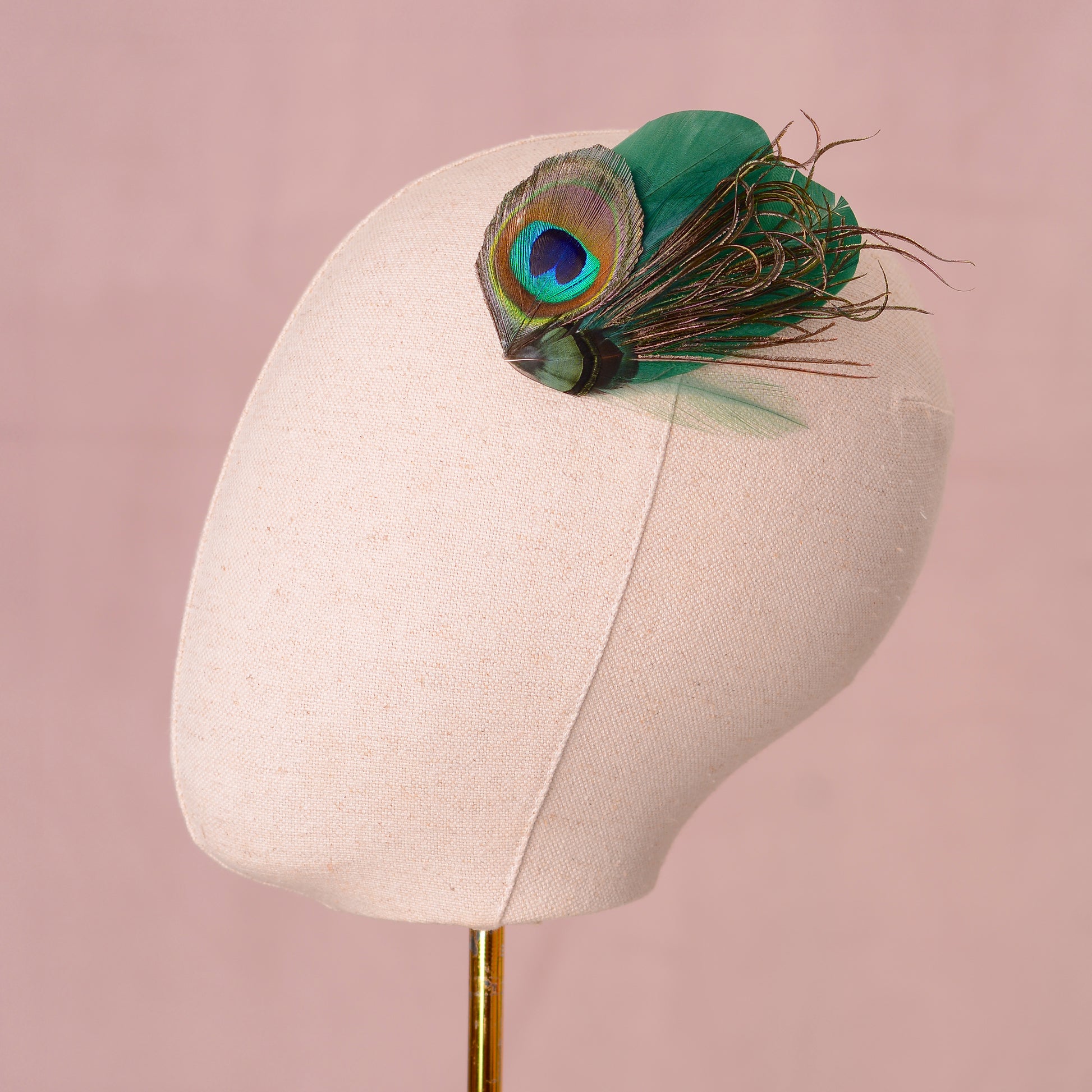 Green peacock feather hair clip on a canvas mannequin head against a pink background