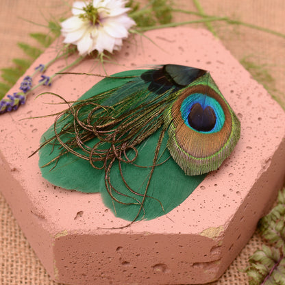 Green peacock feather hair clip on a pink stone with flowers and greenery