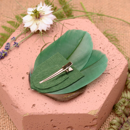 Green peacock feather hair clip on a pink stone with flowers and greenery placed upside down to show clip attachment in a crocodile style