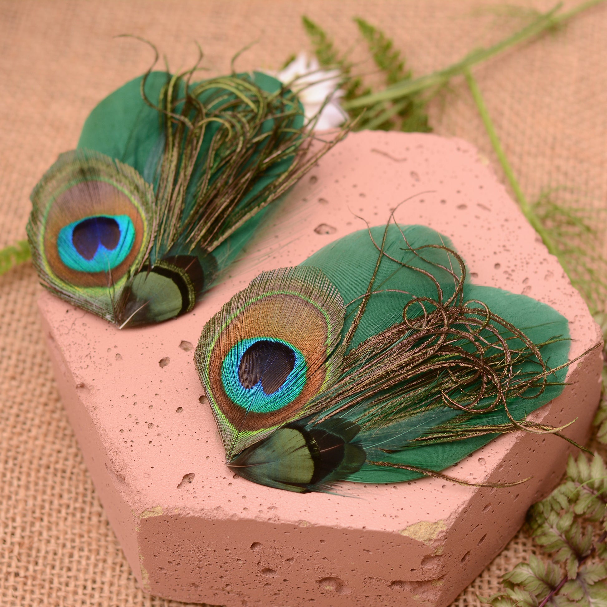 Green peacock feather hair clips on a pink stone with flowers and greenery