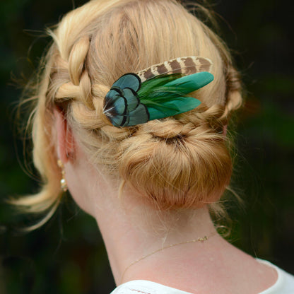 A blonde woman shown from the back wearing a braided bun with a decorative hair accessory featuring green and brown feathers, photographed outdoors against a blurred green background.
