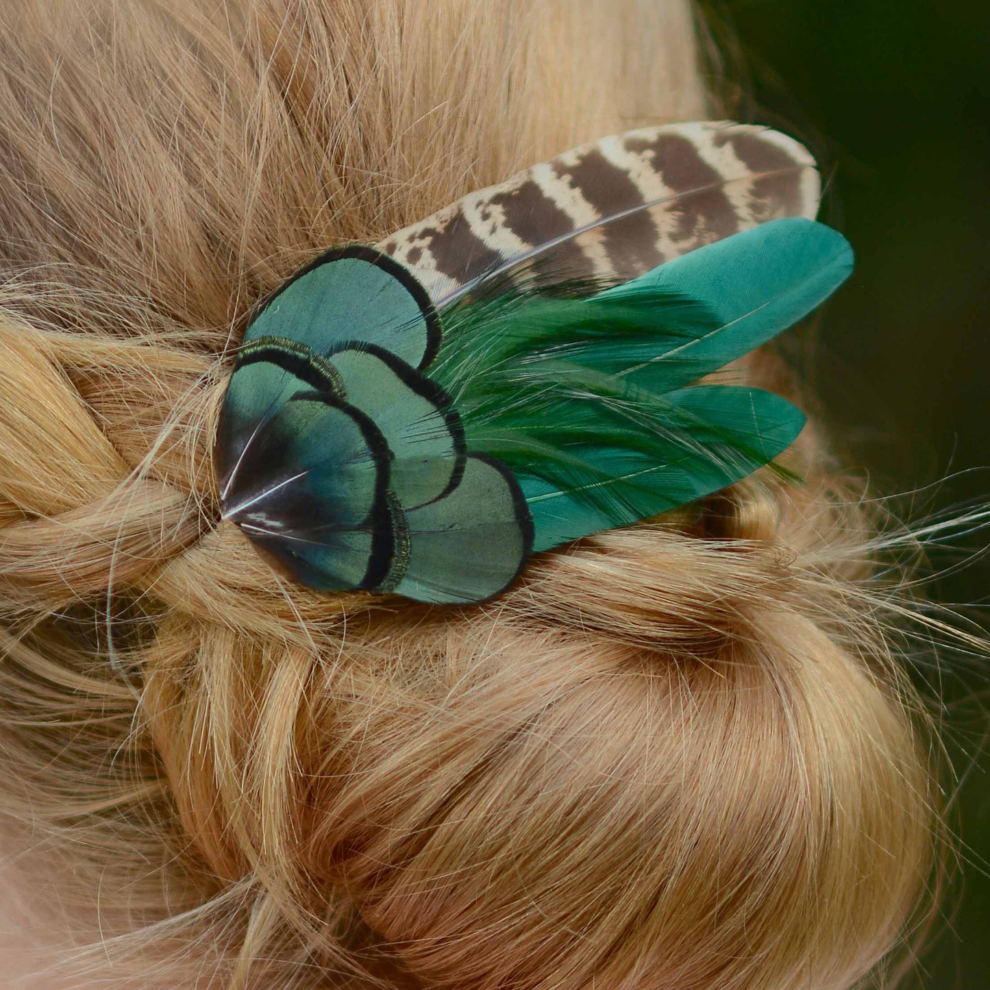 A close-up of a hair accessory clip worn in blonde hair, featuring teal and green feathers with black edging and a brown-and-beige patterned feather.