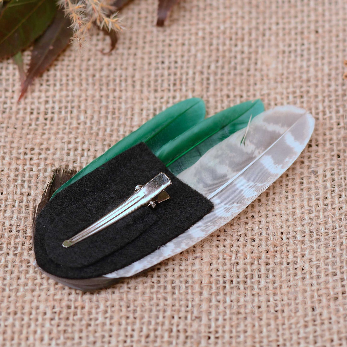 A decorative feather hair clip fascinator. Hand crafted in rich emerald green feathers and brown-and-white banded pheasant feathers.  
The feathers are mounted on a black felt base with a metal alligator style clip. On a textured beige burlap background with dried leaves as props. 