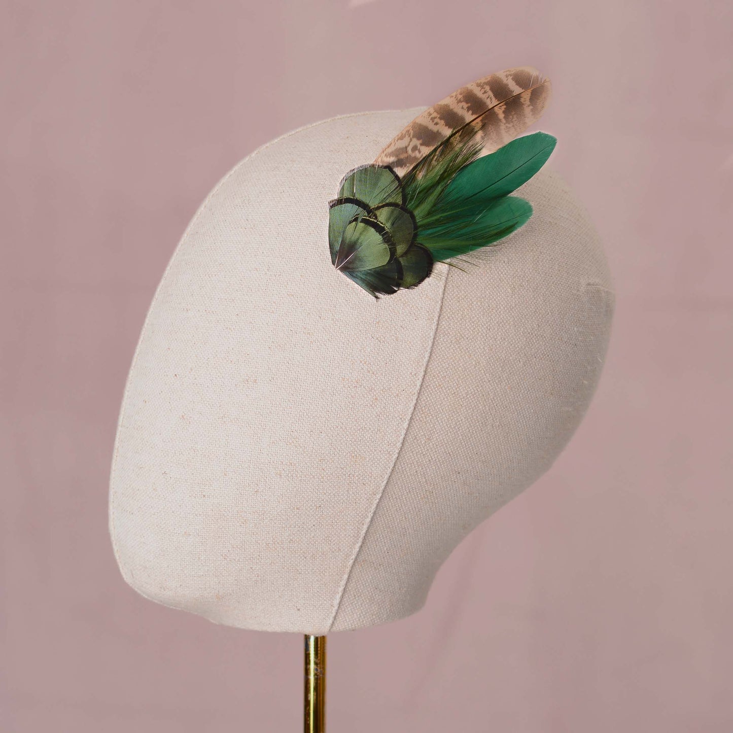 A decorative feather hair accessory with iridescent green and a blonde / brown feather, displayed on a cream fabric mannequin head against a soft pink background.