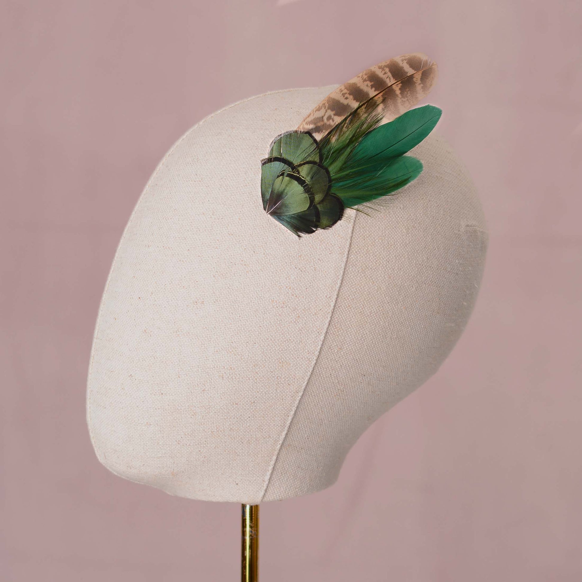 A decorative feather hair accessory with iridescent green and a blonde / brown feather, displayed on a cream fabric mannequin head against a soft pink background.