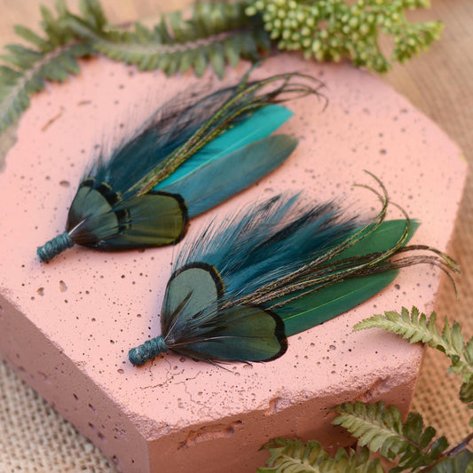 Two handmade feather pins with dark teal, green, and turquoise feathers, displayed on a textured pink hexagonal block with green fern leaves.