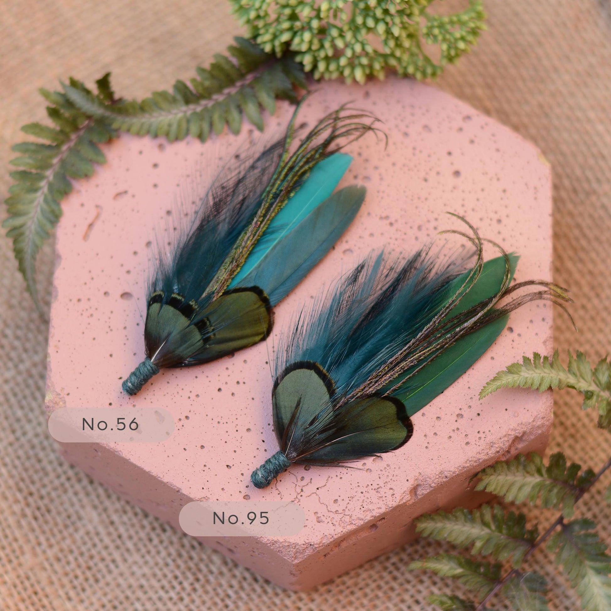 Two feather boutonnieres displayed on a pink stone slab with fern leaves around them. Each feather has dark green and teal-blue feathers with iridescent green edges is tied with teal twine.
