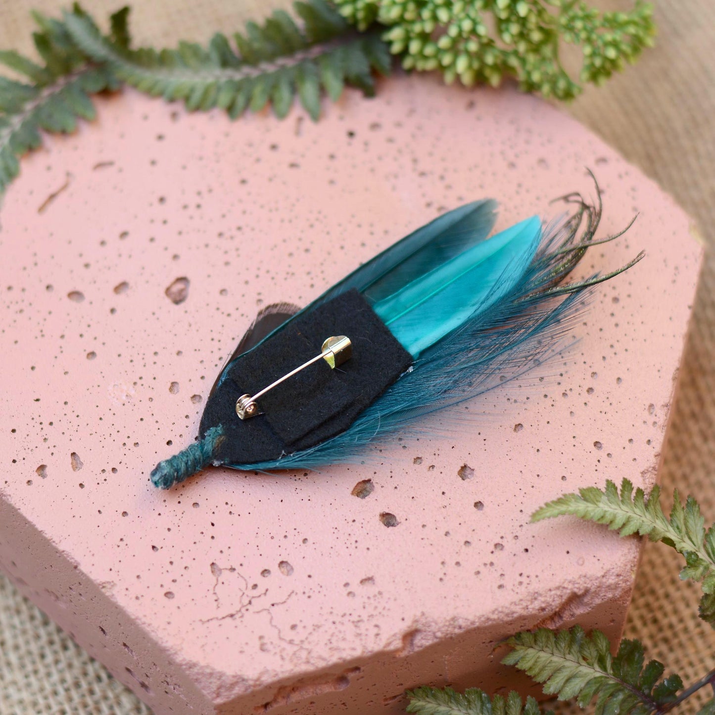 A decorative brooch featuring teal and turquoise feathers attached to a black felt base with a silver-tone brooch pin, displayed on a pink textured stone block with green fern leaves.