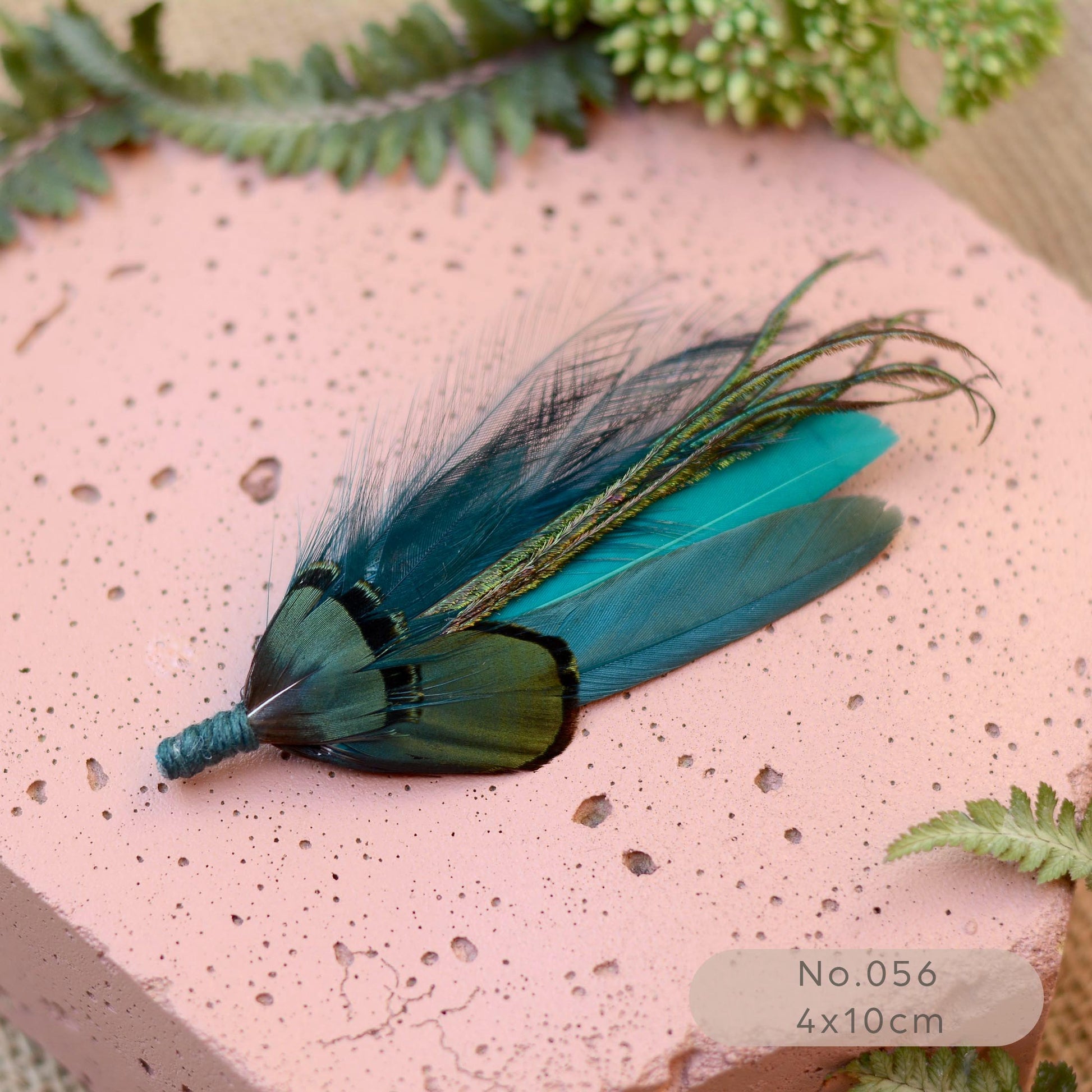 A decorative feather accessory with iridescent teal, blue, and green feathers, displayed on a textured pink surface with fern leaves around it. 