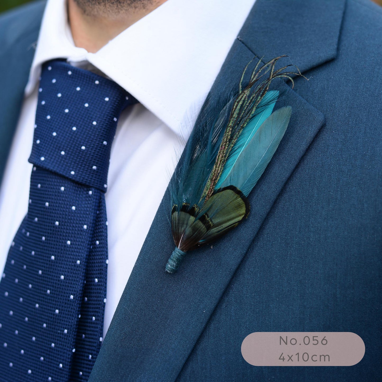 A close-up of a men's formal outfit featuring a blue suit jacket, a navy blue polka dot tie, and a white dress shirt. A decorative feather boutonniere with teal, green.