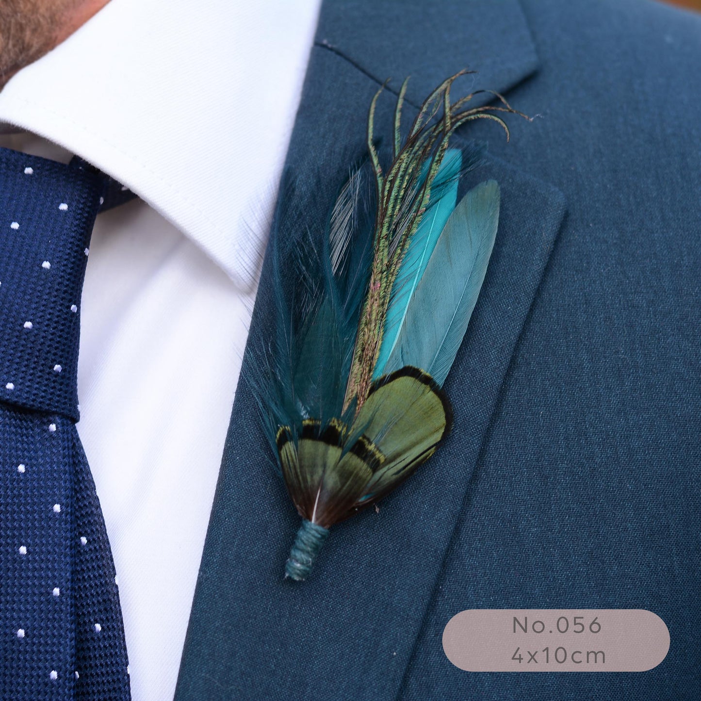 A close-up of a men's necktie and suit jacket with a decorative feathered grooms boutonniere pinned to the lapel. The tie is navy blue with white polka dots, the shirt is white, and the jacket is dark blue.