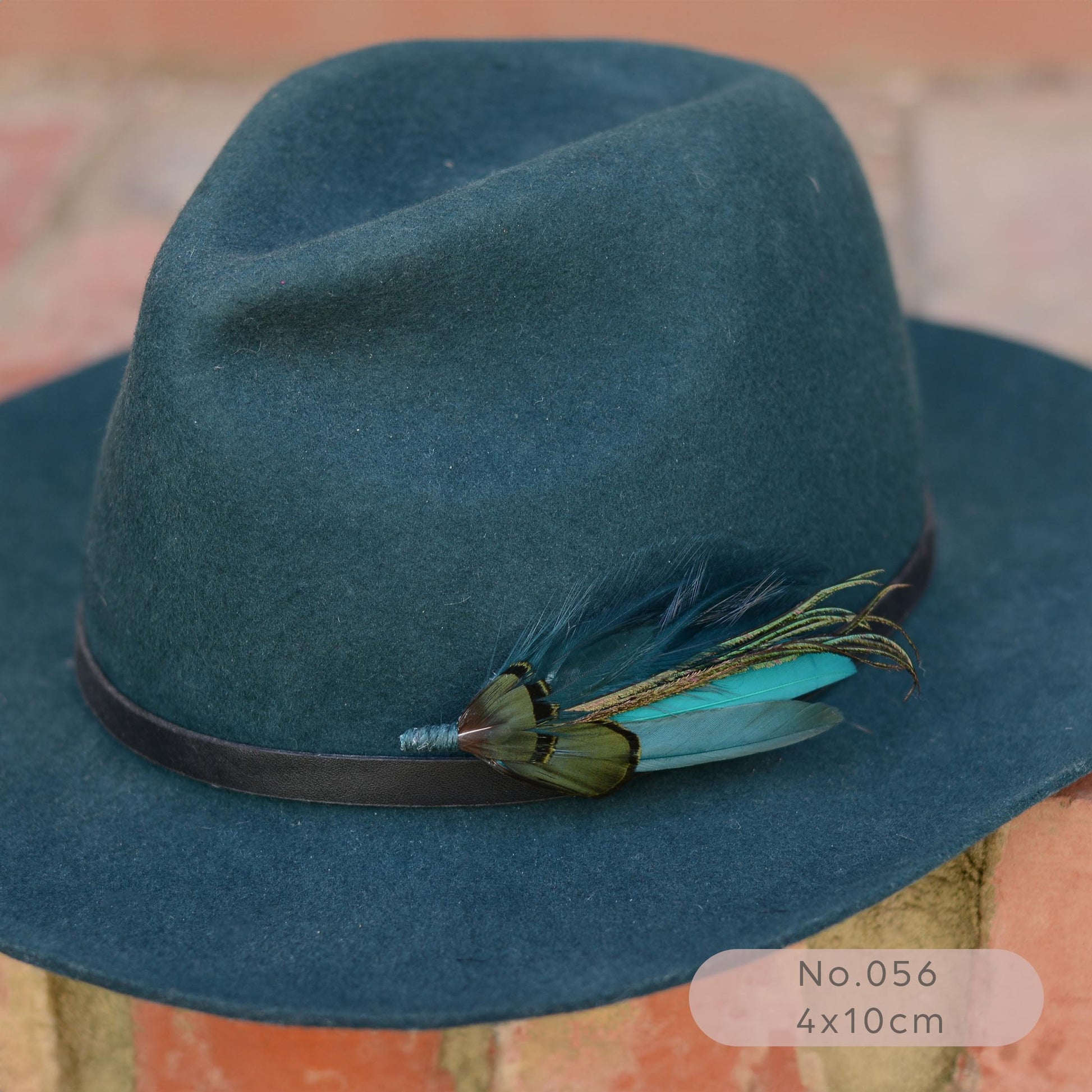 A dark teal felt fedora hat with a black band and a decorative feather brooch pin in teal and green resting on a brick surface.