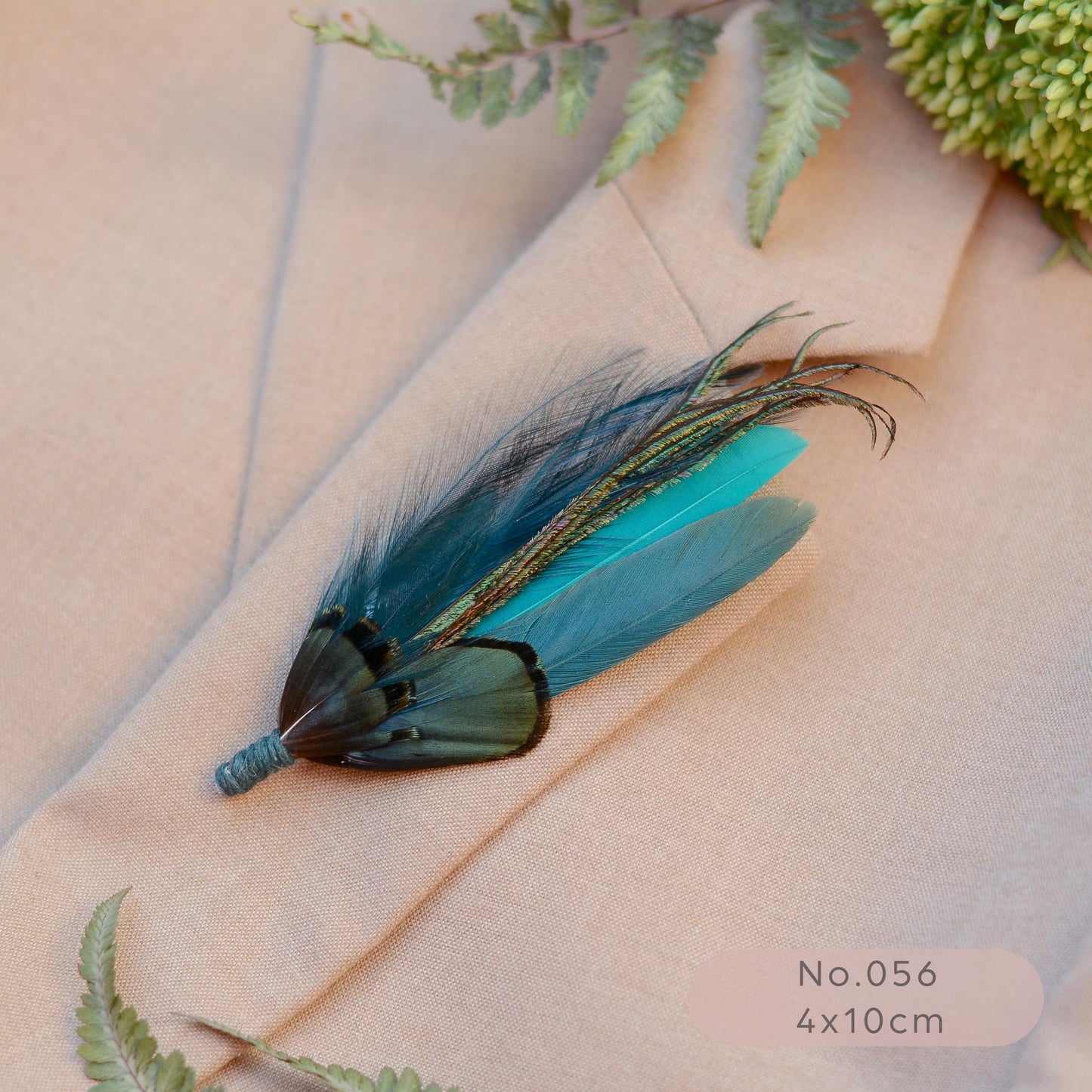 A decorative feather lapel pin  with turquoise and teal feathers lies on a beige suit jacket lapel, with fern leaves and a green floral sprig around it. 