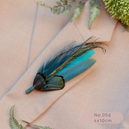 A decorative feather lapel pin  with turquoise and teal feathers lies on a beige suit jacket lapel, with fern leaves and a green floral sprig around it. 