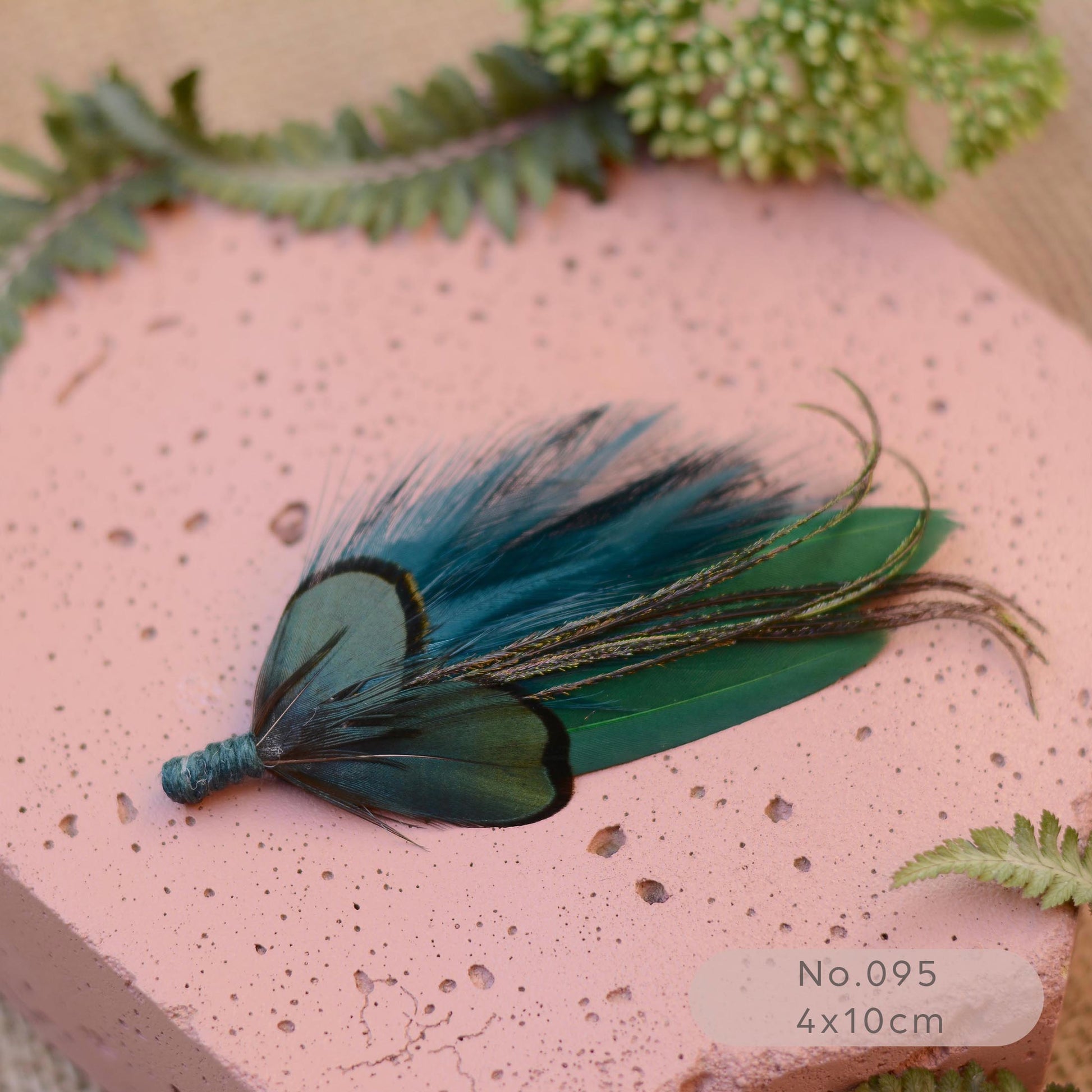 A decorative feather buttonhole brooch with teal, blue, and green feathers lies on a pink speckled stone slab, with green foliage around it. 