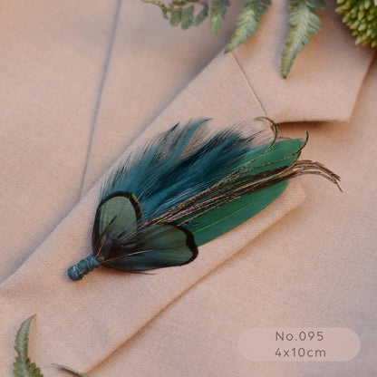 A decorative feather boutonniere brooch with teal and green feathers tied with teal  blue cord, laid on a beige suit jacket with fern leaves around it. 