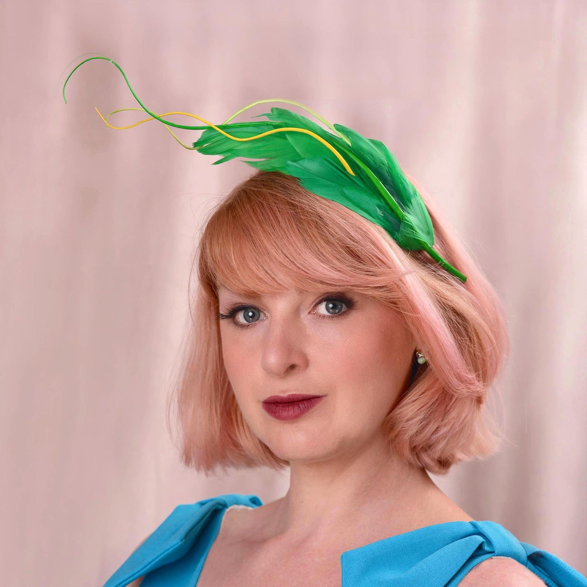 A woman with short pink hair wearing a green feather fascinator headpiece and a bright blue top, photographed against a soft beige curtain background.