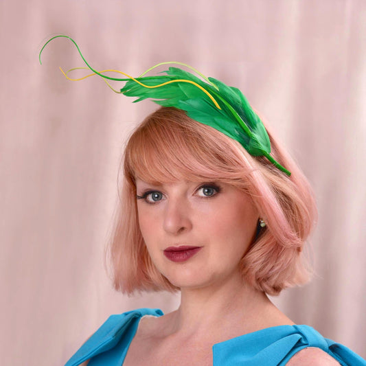 A woman with short pink hair wearing a green feather fascinator headpiece and a bright blue top, photographed against a soft beige curtain background.