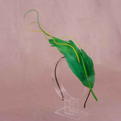 A green feather fascinator headpiece with yellow and green accents, displayed on a clear acrylic stand against a dusty pink background.