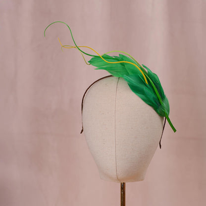 A green and yellow feather fascinator headpiece displayed on a cream fabric mannequin head against a soft pink background.