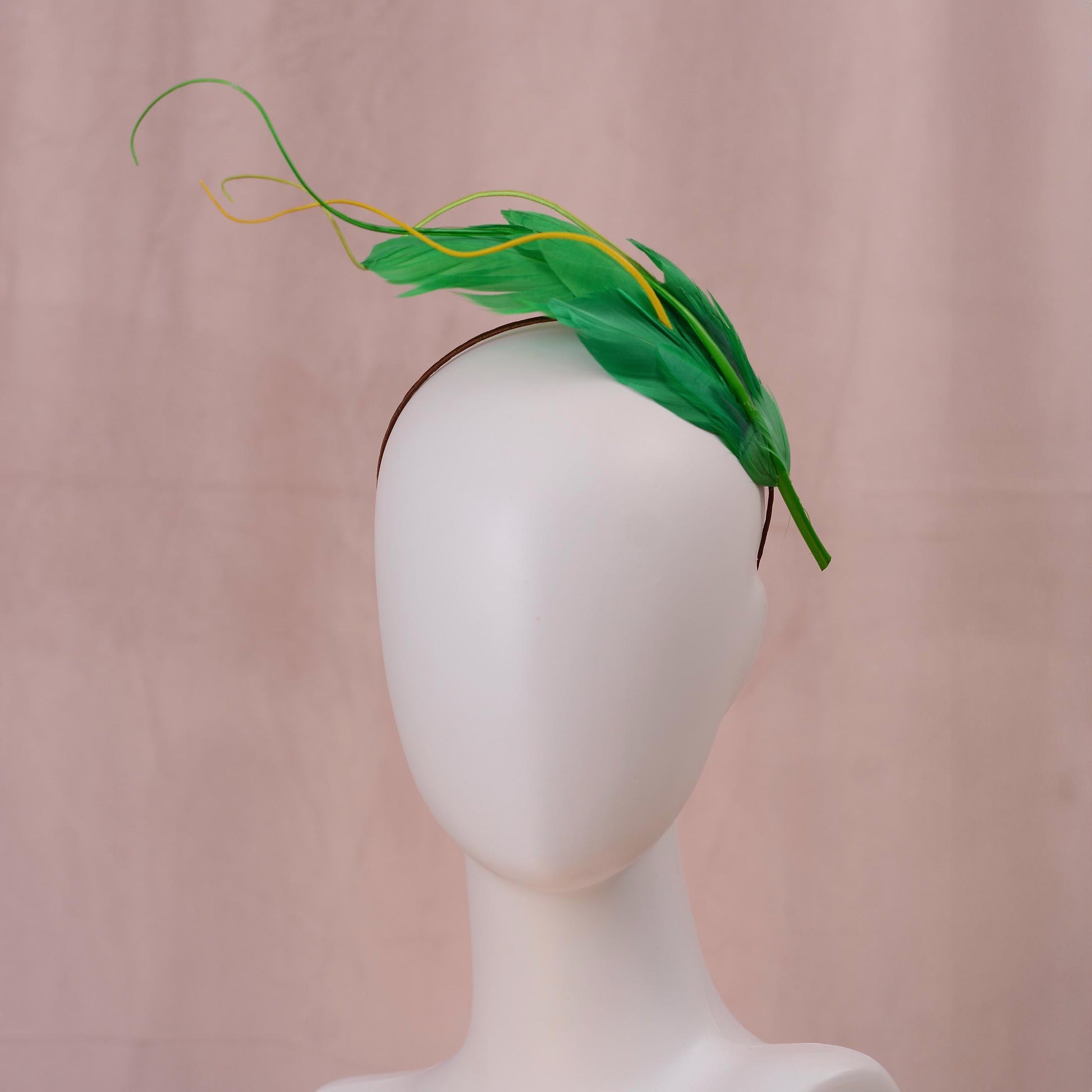 A green floating feather  fascinator headpiece with yellow feather accents, displayed on a white mannequin head against a soft pink backdrop.