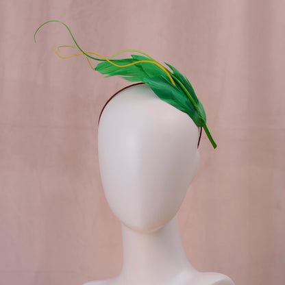 A green floating feather  fascinator headpiece with yellow feather accents, displayed on a white mannequin head against a soft pink backdrop.