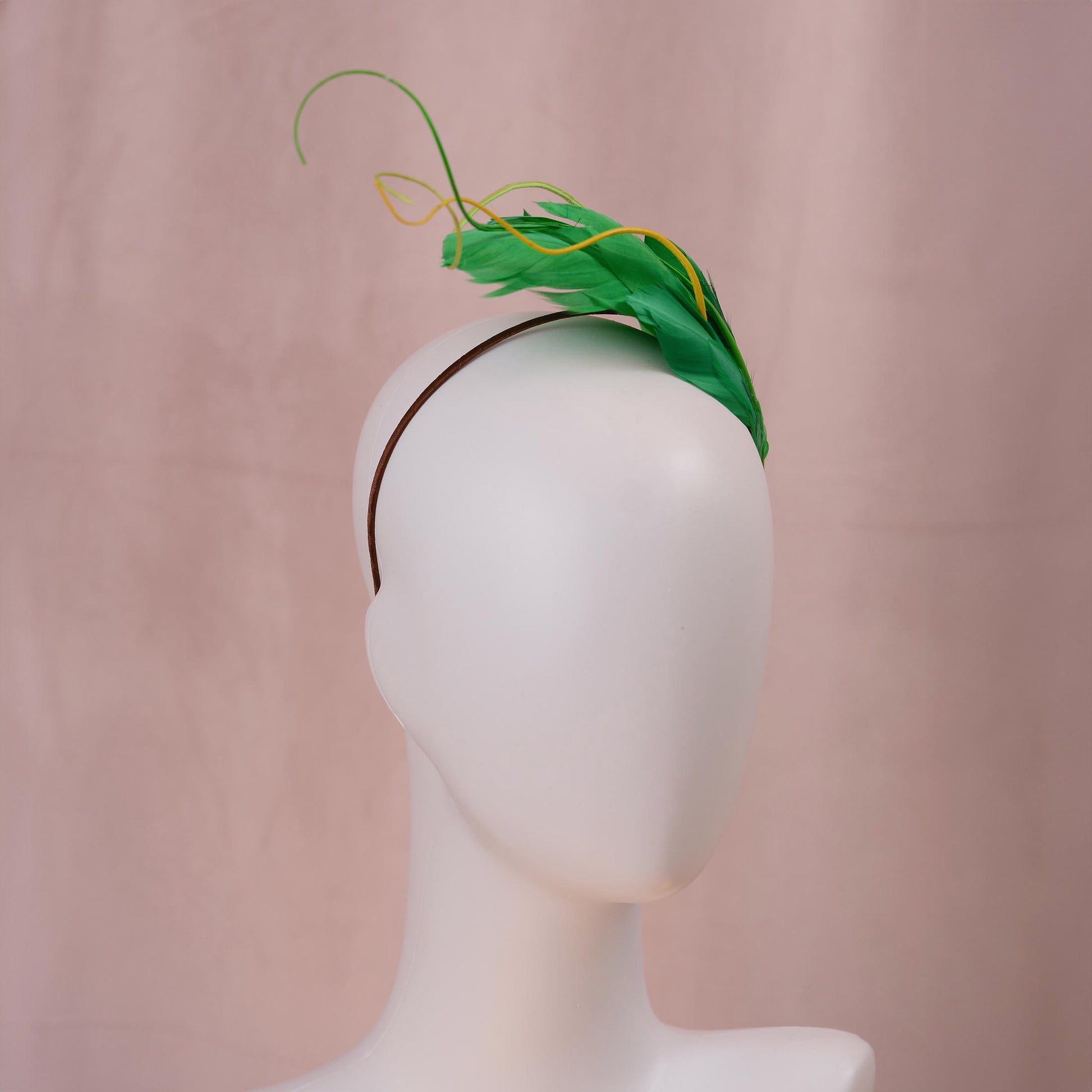 Close up of a bright green feather headband with yellow feather accents, displayed on a white mannequin head against a soft pink backdrop.