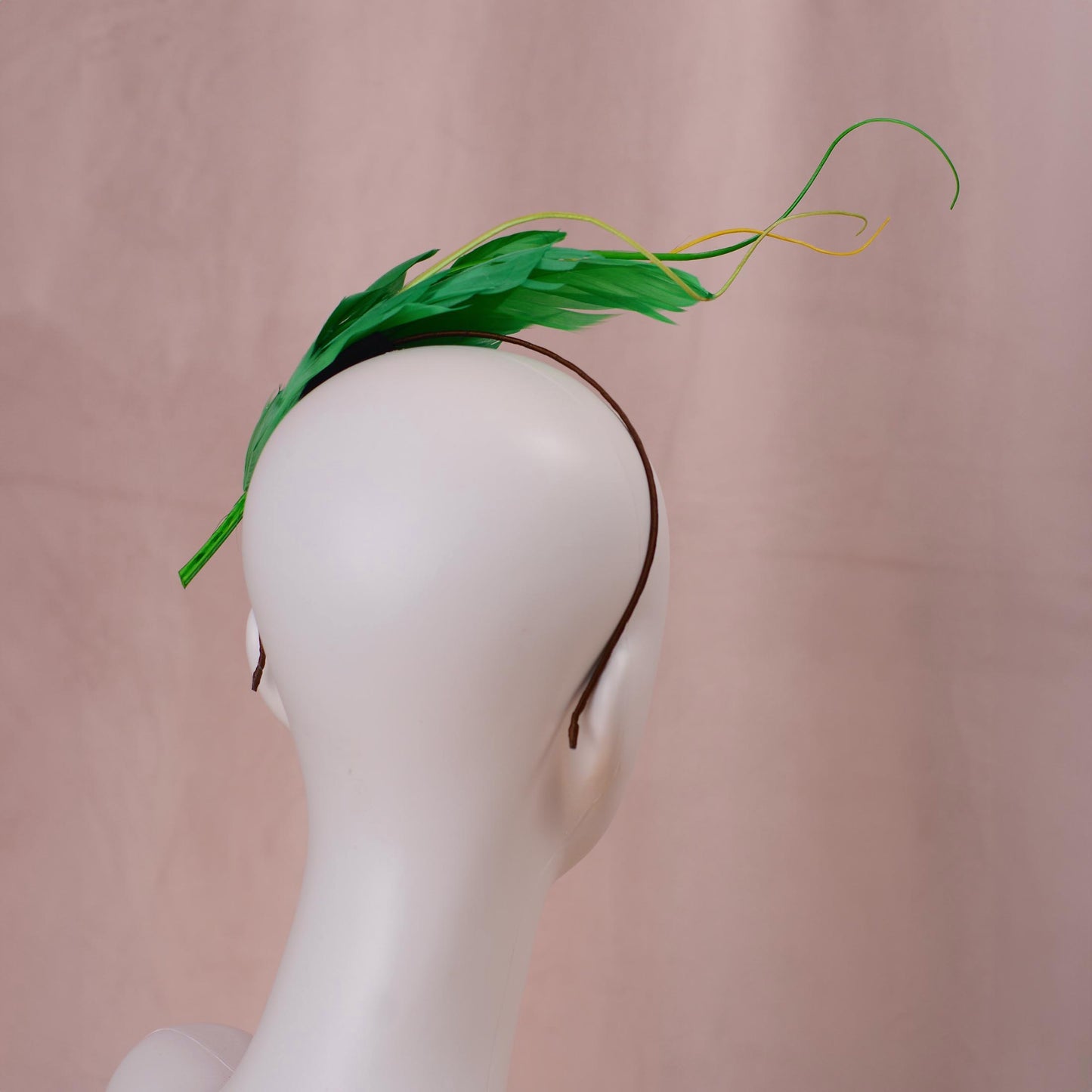 A green feather fascinator headpiece with a thin brown headband, displayed on a white mannequin head against a soft pink background.
