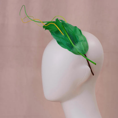 A green floating feather wedding headband with yellow feather accents, displayed on a white mannequin head against a soft pink backdrop.