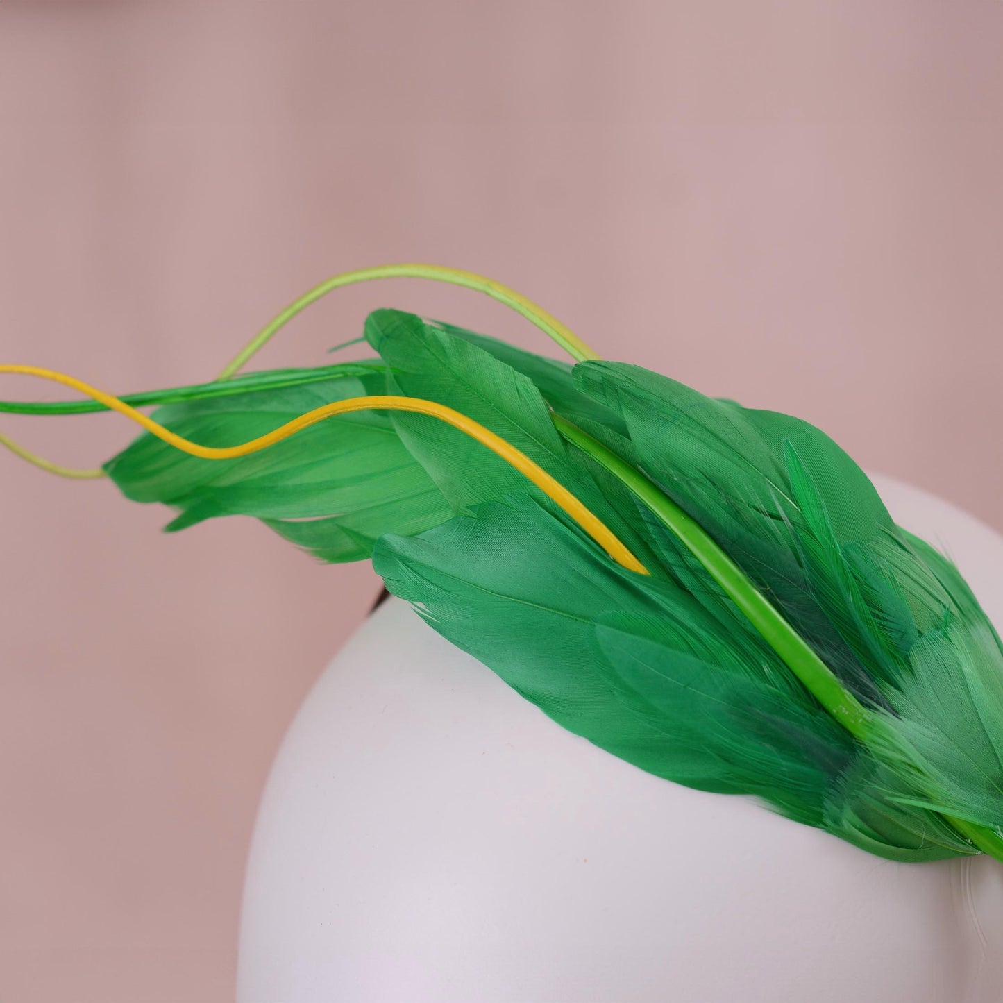 A green feather fascinator headpiece with a thin brown headband, displayed on a white mannequin head against a soft pink background.