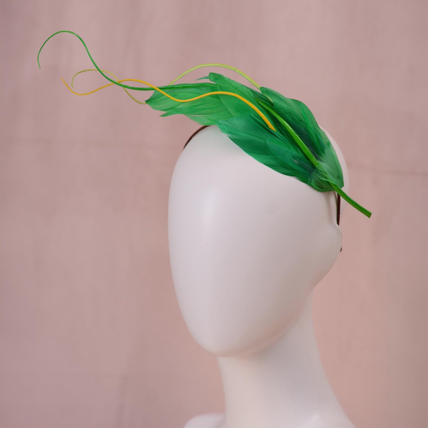 A green floating feather wedding headpiece with yellow feather accents, displayed on a white mannequin head against a soft pink backdrop.