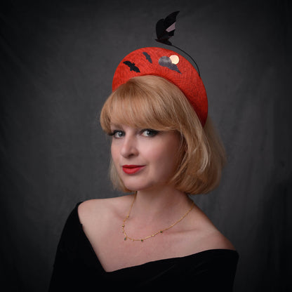 A woman with a short blonde bob wears a bright red fascinator hat decorated with black bat silhouettes, a cloud, and a metallic pink blood moon with a floating bat, posed against a grey background.
a headpiece for Halloween-themed weddings, events and parties