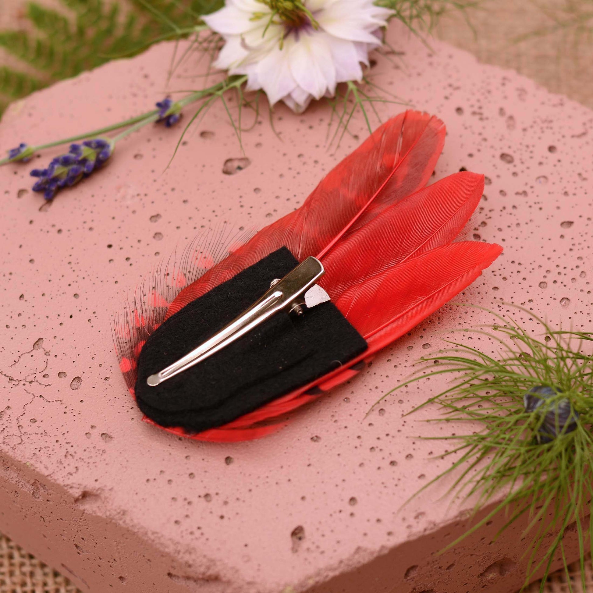 A red feather hair clip shown upside down to view the felt base and alligator style hair attachment