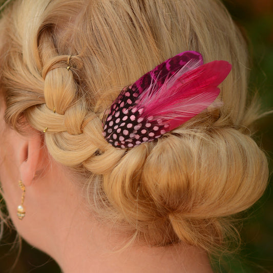 Blonde hair styled in a braid with a pink and black feather accessory, blurred green background