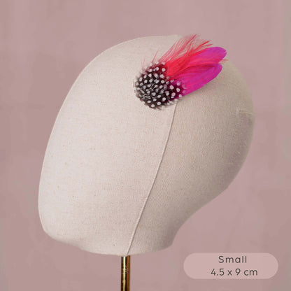 A small feather hair accessory displayed on a cream fabric mannequin head against a dusty pink background. The accessory features black and white spotted feathers and bright pink feathers.