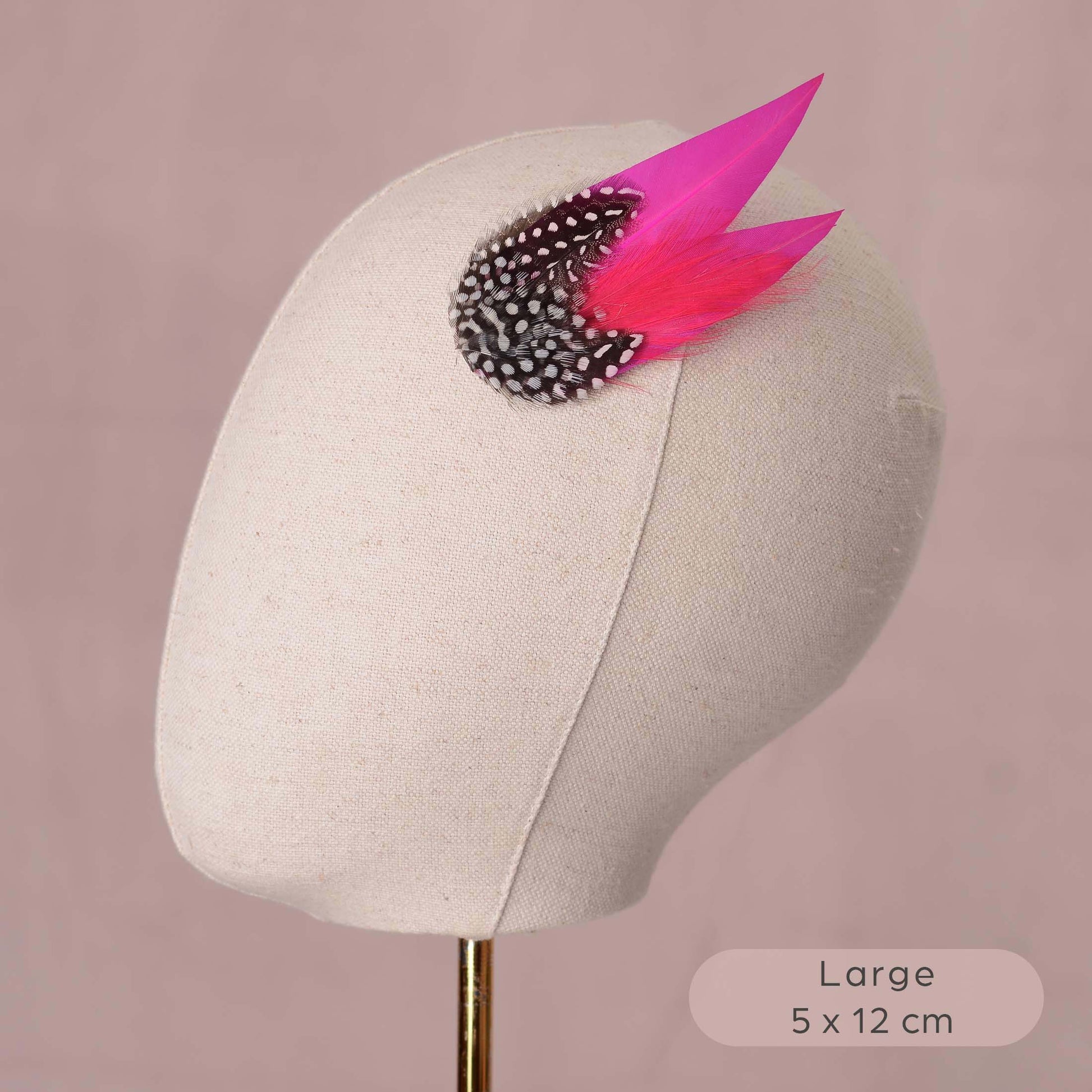 A decorative hair accessory with two bright pink feathers and a black-and-white spotted feathers, displayed on a beige fabric mannequin head against a dusty pink background. A text label in the bottom right reads 'Large 5 x 12 cm'.
